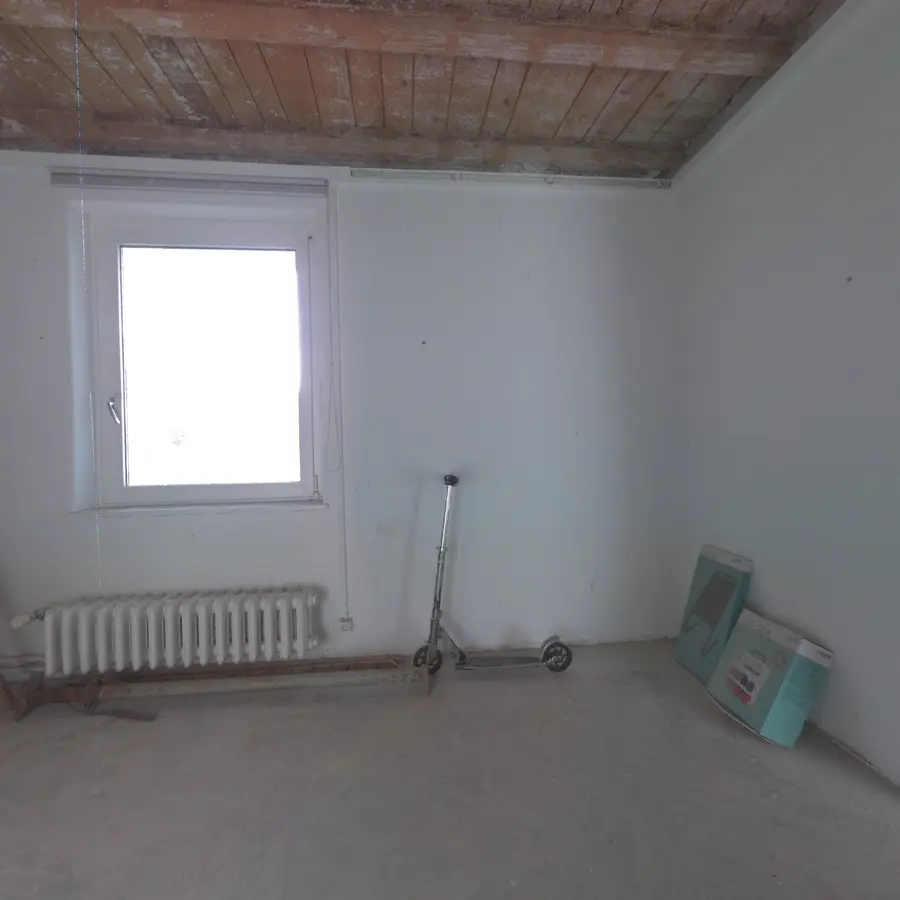 An empty room photographed from the doorway: window, radiator, a scooter against the wall.